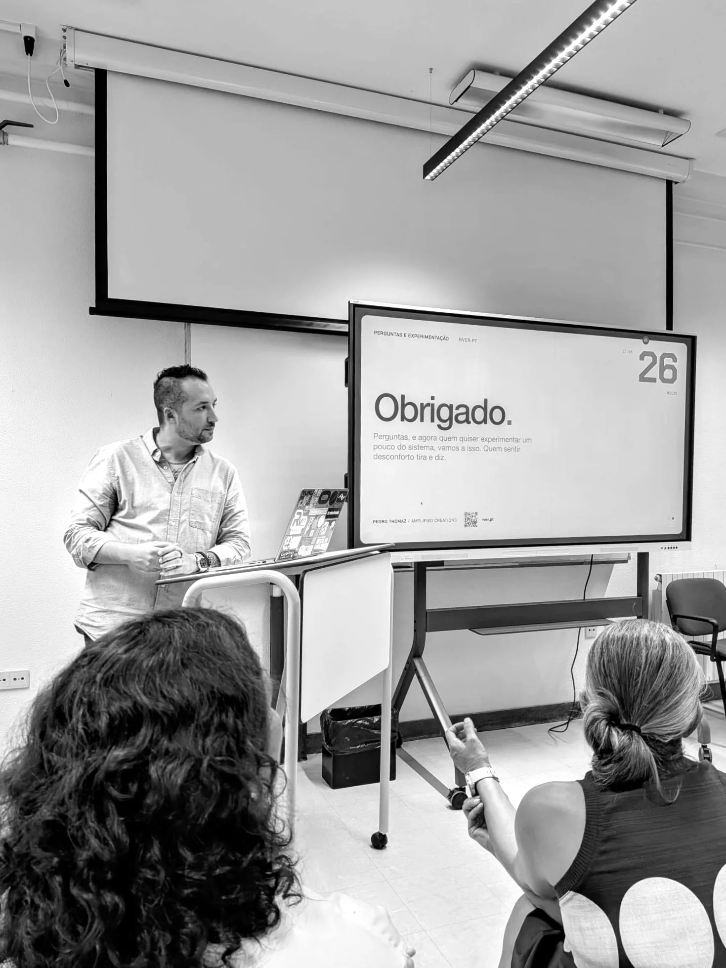 A classroom with a large screen reading «Obrigado», and two people in the audience seen from behind.