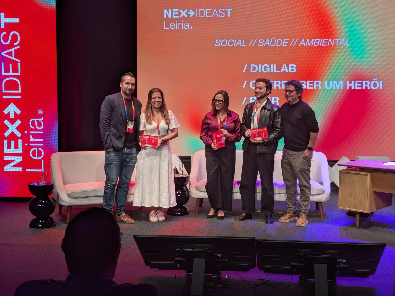 Os quatro oradores do painel e o Fernando Alvim de pé no palco; três seguram uma peça vermelha do NEXXT Ideas.