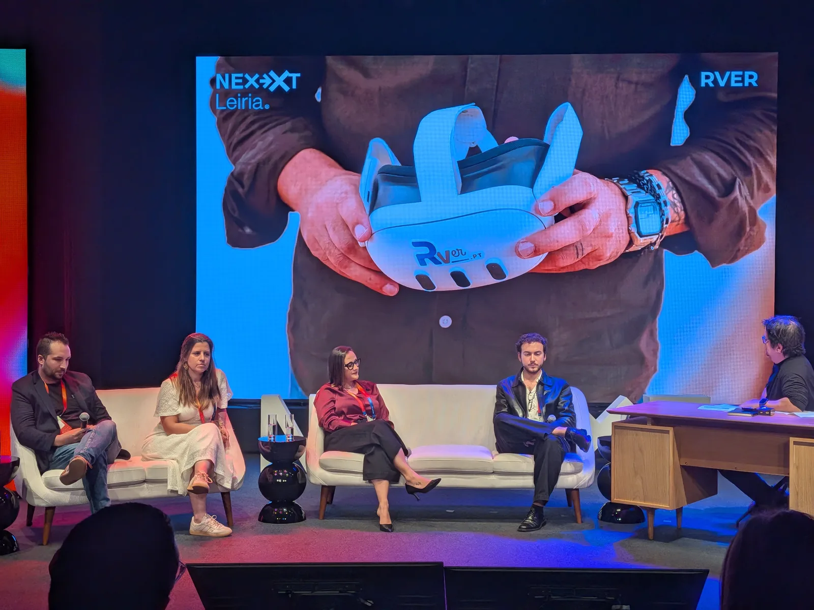 Palco do NEXXT Ideas com quatro oradores sentados em sofás brancos e um moderador à secretária; no ecrã grande, duas mãos seguram um headset com o logótipo RVer.