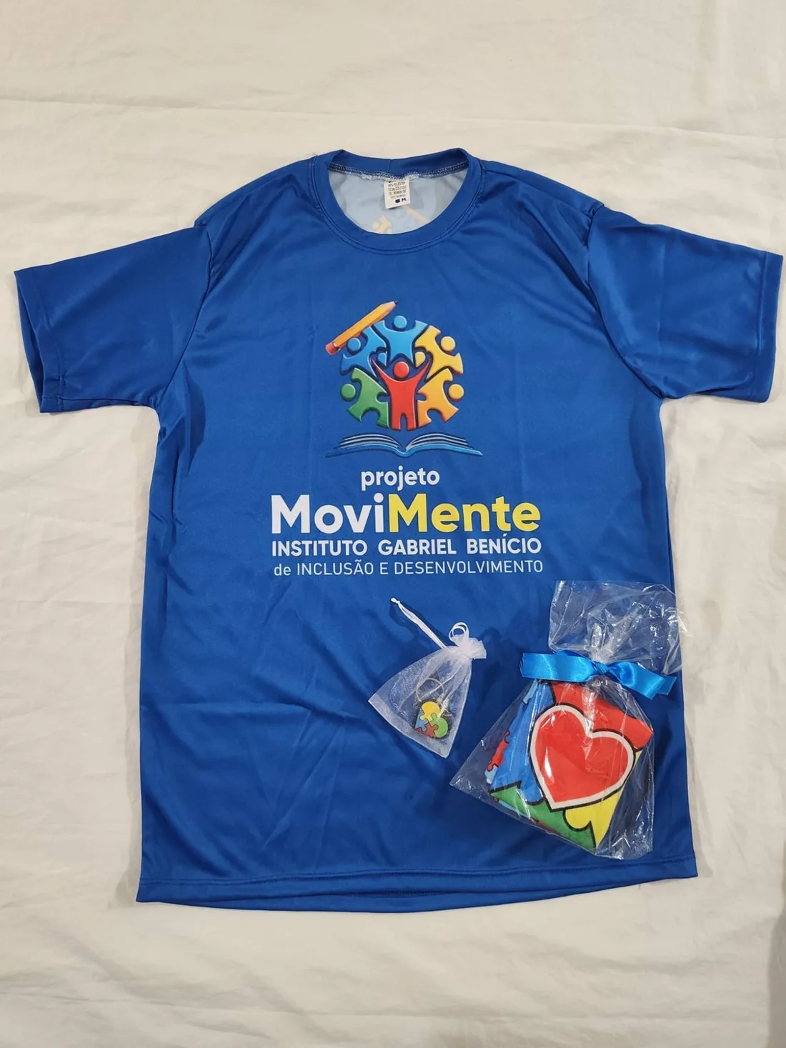 Camisola azul do projeto MoviMente, do Instituto Gabriel Benício de Inclusão e Desenvolvimento, com o logótipo em peças de puzzle coloridas, um lápis e um livro aberto.